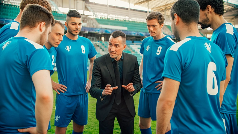 Imagem mostra um técnico conversando com os jogadores de futebol em campo