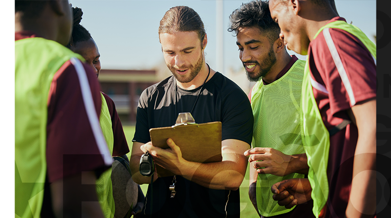 Imagem mostra jogadores em campo analisando informações numa prancheta com o técnico